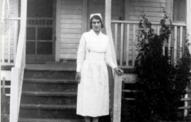 A young woman dressed as a nurse is standing on the front steps of a wooden house.