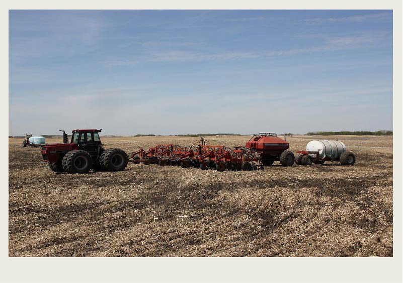 A tractor pulls an air drill, a red tank with seed, and a white tank with fertilizer on a field to seed the crop.