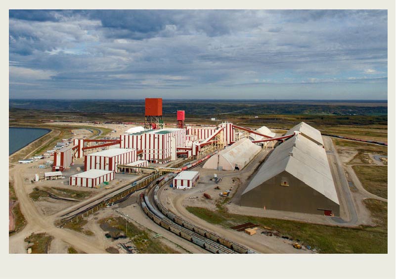 An aerial view of a Saskatchewan potash mine with many large buildings and track tracks coming up to the buildings is shown with a cloudy sky over it.