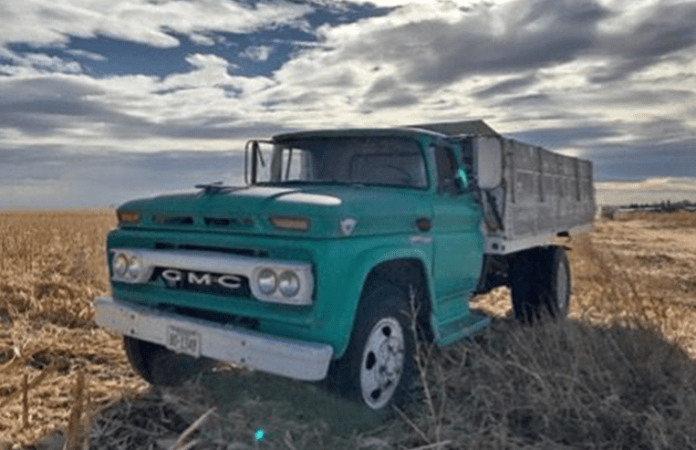 A blue grain truck is parked in a farm field with a bright sky above.