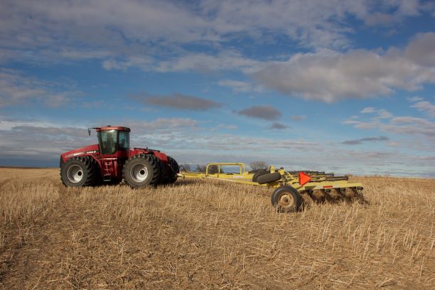 A tractor with tracks pulls a large set of harrows across a farm field.