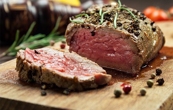 A elk roast is sitting on a wooden platter with peppers and spices on it.