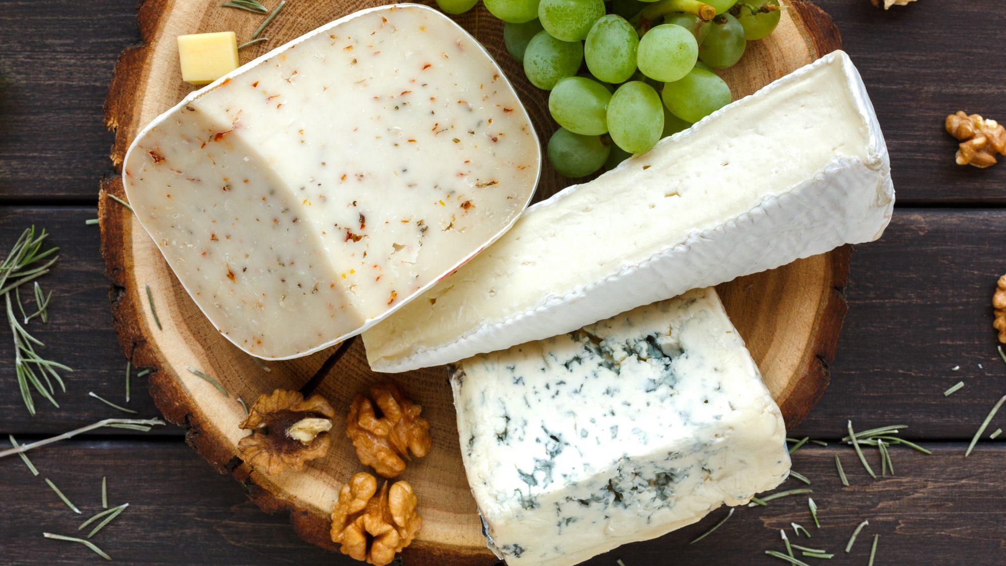 A wooden bowl with grapes and a variety of goat cheeses sits on a wooden table.