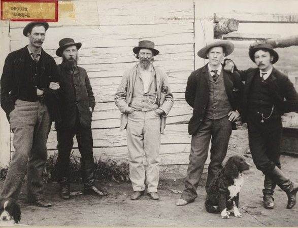 A group of men stand together outside of a wooden building in Fort Pitt in 1884.  Left to right: Thomas Trueman Quinn, Indian Agent; Inspector Francis Jeffries Dickens, NWMP, son of the novelist, in command of the Fort Pitt Detachment; James Keith Simpson, son of Sir George Simpson, the great Governor of the Hudson's Bay Company; Frederick Stanley Simpson, HBC clerk, Fort Pitt; Angus McKay, Post Manager, HBC, Fort Pitt.