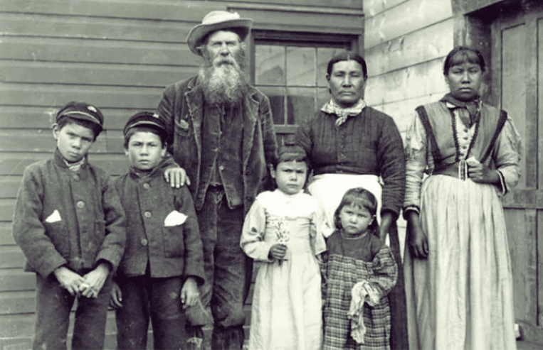 A European man, two First Nation women, and four Métis children are standing in front of a wooden house.