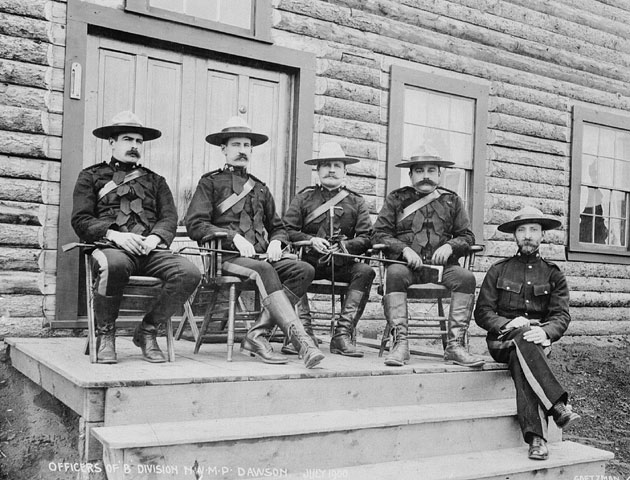 Five North-West Mounted Police are wearing their uniforms and sitting on the front deck of a log building. 