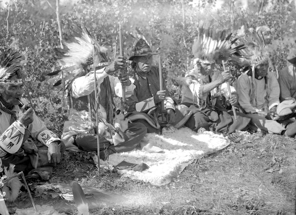 A group of Cree men sit in a circle in traditional clothing. 