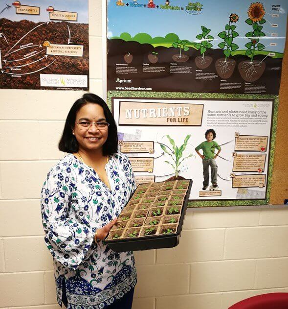 A woman holds small plants as she stands next to a poster about growing plants.