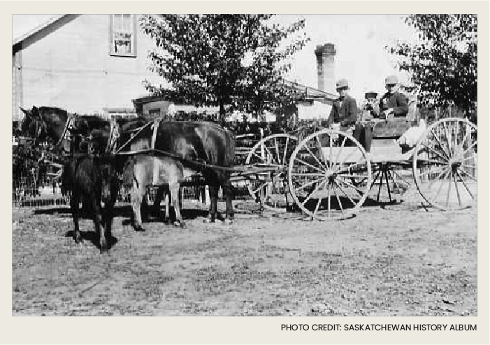 Three young children are in a wooden wagon that is being pulled by a team of two horses. There's a wooden house in the background.