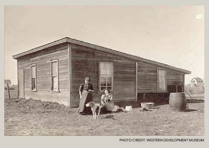 Two women and a dog are standing in front of a wooden house on a homestead.