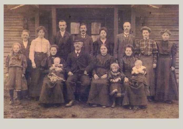 A group of sixteen people have gathered for a community picnic and they are standing for a photo in front of a wooden building. 