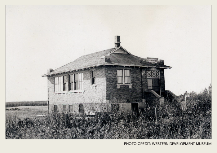 Photo caps-01 copy 2 The outside of this one-room school house is brick and it has a front door with steps that lead down to a grassy field.