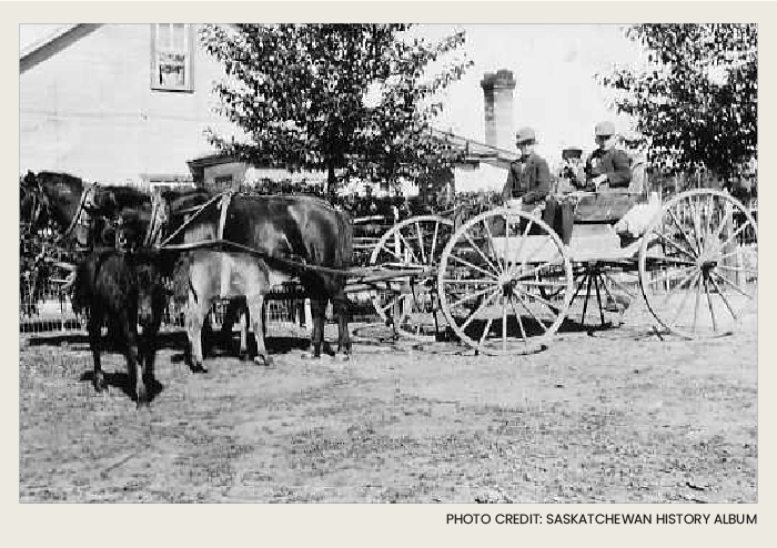 Photo caps-07 copy Three young children are in a wooden wagon that is being pulled by a team of two horses. There's a wooden house in the background.