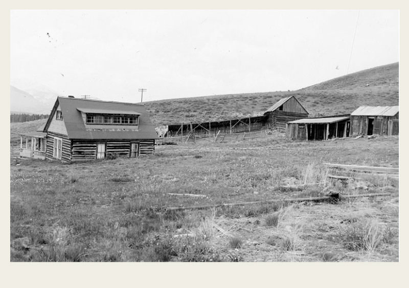 A ranch with a house and barn and fences stand in the bottom of a green-grass valley. 