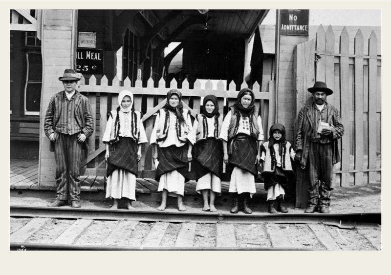 A Ukrainian family is dressed in traditional outfits and lined up outside a fence by a train station for a photo. 