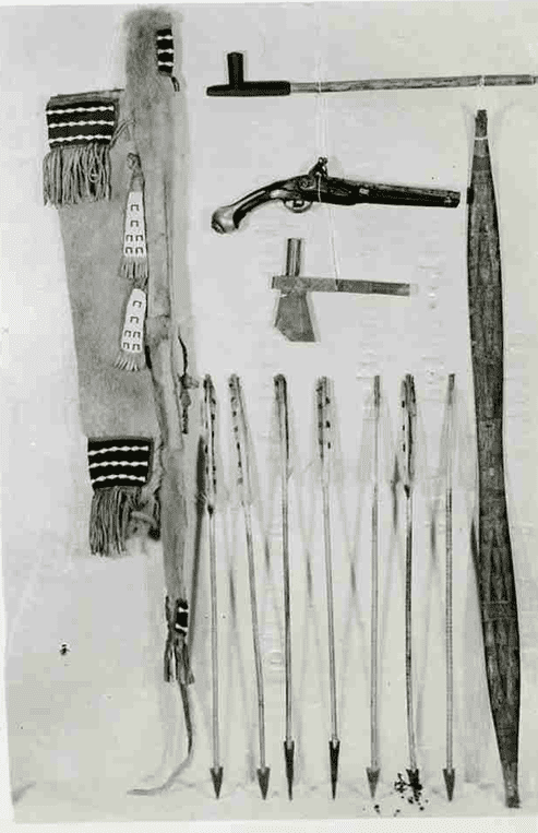 A black and white photograph of various Aboriginal artifacts including a bow and quiver with arrows, a flintlock pistol, an axe head, and a pipe.